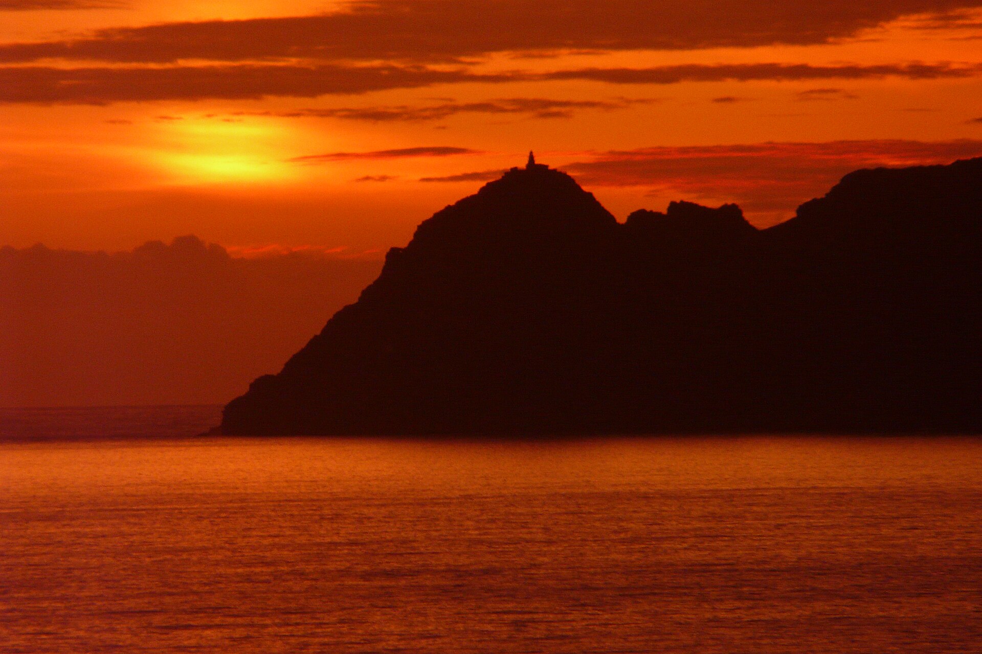 Silueta de las Illas Cíes al atardecer, faro en el pico y mar en calma bajo un cielo naranja.