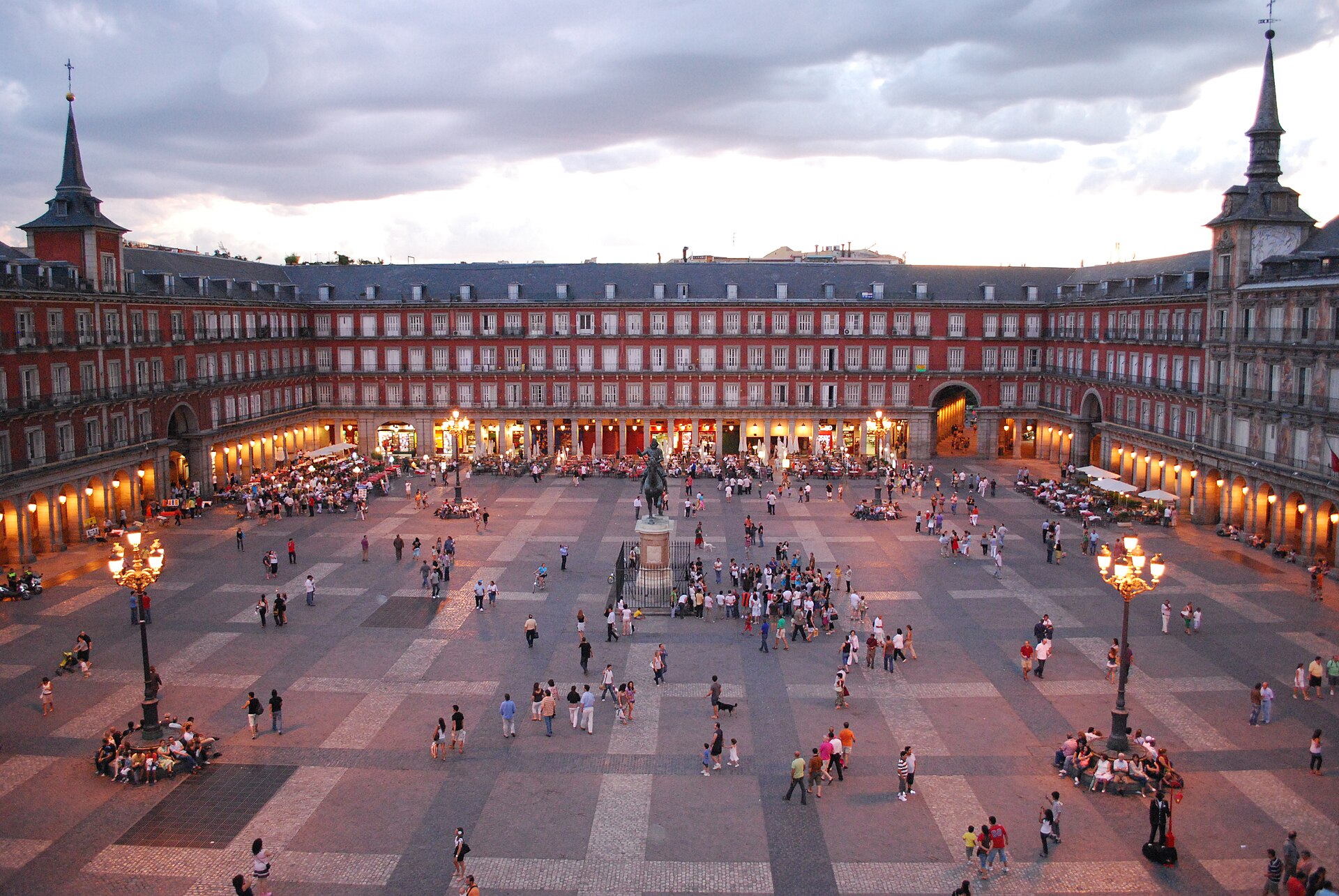 Plaza Mayor de Madrid al anochecer, fachadas rojas, estatua de Felipe III y gente en el enlosado.